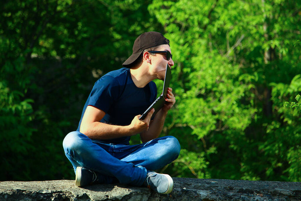Молодий чоловік сидить на бетонному бордюрі в парку і емоційно цілує ноутбук у передній камері
. - Фото, зображення