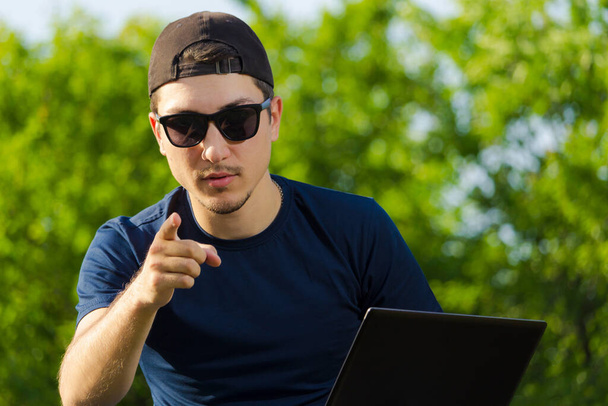 Nuori miespuolinen opiskelija tai freelancer istuu puistossa kannettavan tietokoneen kanssa ja näyttää sormea. Käsite: Voit tehdä sen liian
 - Valokuva, kuva