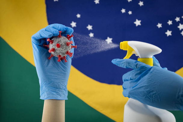 Drewniany model dłoni noszący niebieskie rękawiczki, rozpylający piłeczkę koronawirusową z brazylijską flagą na tle. - Zdjęcie, obraz