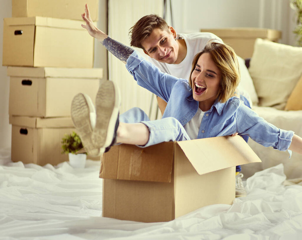 Happy couple having fun and riding in cardboard boxes at new home - 写真・画像