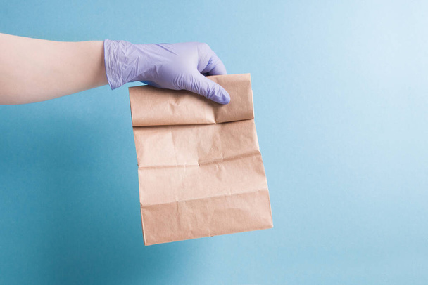 contactless food delivery concept, courier's hand in a blue rubber glove holds an order, paper bag on a blue background, copy space - Photo, Image