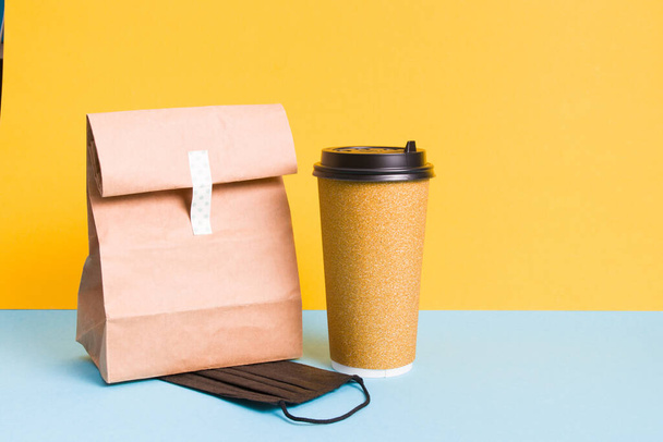 contactless food delivery concept, black protective mask, cardboard cup of coffee in golden shiny packaging and paper bag, light blue and yellow background, copy space - Φωτογραφία, εικόνα