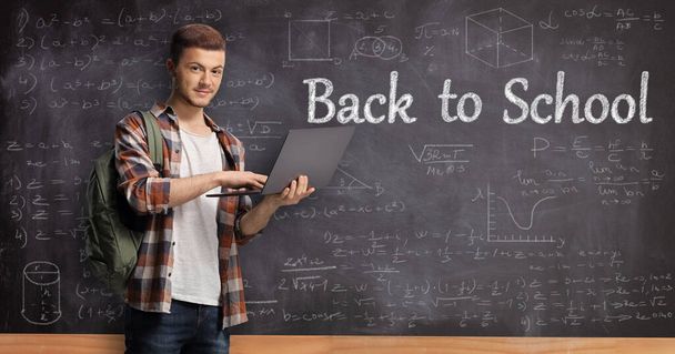 Male student holding a laptop computer in front of a blackboard with formulas and text back to school - Photo, image