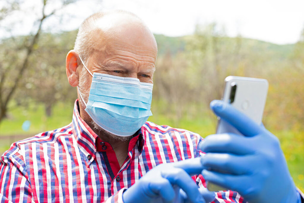 Egy idős férfi portréja, aki sebészeti maszkot és kesztyűt visel, miközben a szabadban telefonál. - Fotó, kép
