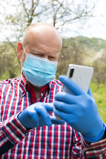 Portrait of elderly man wearing surgical mask and gloves talking on the phone outdoors - Photo, image