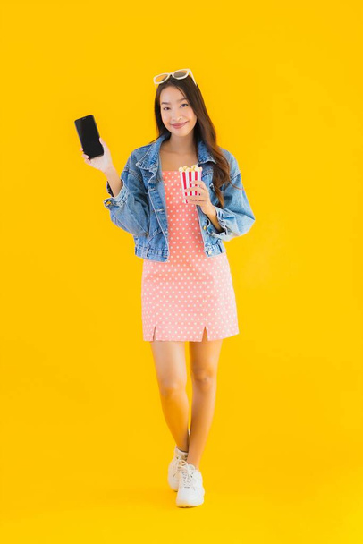 Портрет красивая молодая азиатская женщина наслаждаться счастливым с телефоном попкорн и смотреть фильм на желтом изолированном фоне
 - Фото, изображение