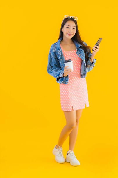 Portrait beautiful young asian woman with coffee cup and smart mobile phone on yellow isolated background - 写真・画像