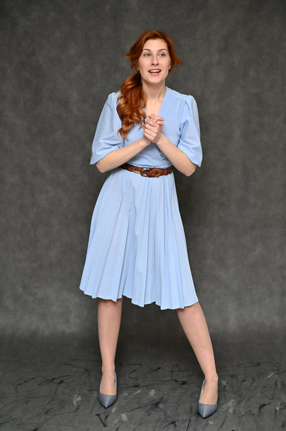 Model demonstrates standing different poses. Full-length Vertical Portrait of a young pretty red-haired woman in a blue dress on a gray background. - Foto, Bild