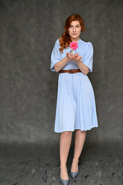 The model demonstrates standing in full growth different poses. Vertical portrait of a young pretty red-haired woman in a blue dress on a gray background. - Foto, Bild