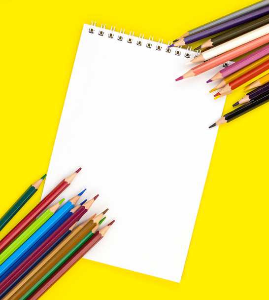 white blank Notepad in the center and colored pencils in the corners, isolated on a yellow background. stationery for school preparation. background for designers with space for text. online learning - Photo, image