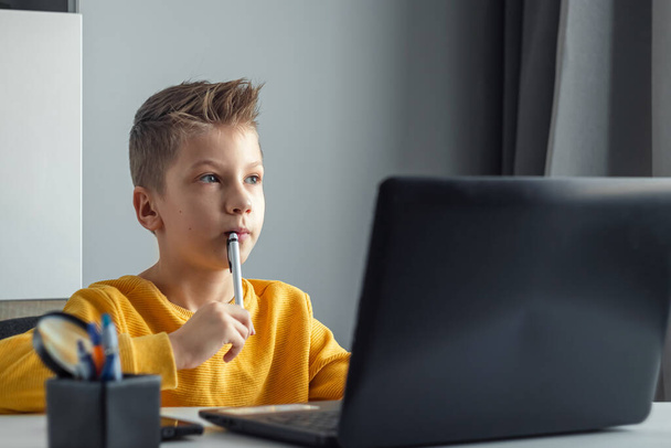 A child, a boy in a yellow jacket, is sitting at a table at home looking at a laptop, online learning, distance learning at home via the Internet. Technology, school, knowledge. Copy space - Fotografie, Obrázek