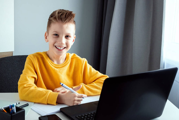 A child, a boy in a yellow jacket, is sitting at a table at home looking at a laptop, online learning, distance learning at home via the Internet. Technology, school, knowledge. Copy space - Fotó, kép
