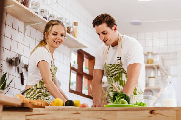 Happy & smiling attractive young cute caucasian couple in love enjoying cooking healthy salad in kitchen at home together. Happiness family of beautiful romantic married lover or marriage relationship - Foto, Imagen