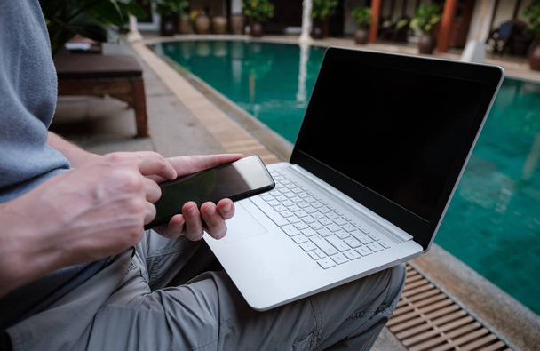 seyahat blogcusu yüzme havuzunda beyaz dizüstü bilgisayar ve akıllı telefonla oturuyor, internet üzerinden seyahat ediyor. Serbest kumanda konsepti. Boşluğu kopyala, deniz manzarası arkaplanı - Fotoğraf, Görsel
