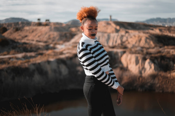 arfican donna nera americana indossa un maglione strip bianco e nero con collo alto, in mezzo al campo e al lago durante il tramonto
  - Foto, immagini