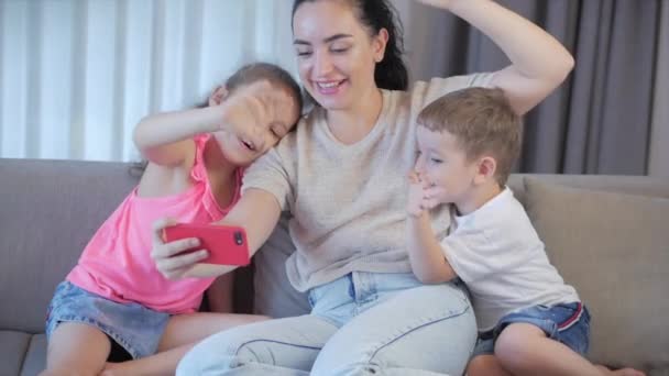 Familia feliz, mamá y lindos niños pequeños, mamá juega con los niños en casa, relajante, usando un teléfono inteligente, linda familia hablando con la webcam haciendo videollamada en línea, sentado en el sofá
. - Metraje, vídeo