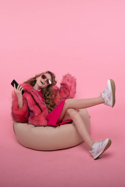 Beautiful young girl sits in a chair a beige bag and sunglasses with a phone in her hands and listens to music. - Photo, Image
