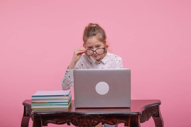 On the table is a laptop and books. A beautiful young girl in glasses for vision sits at a table. - Foto, Bild