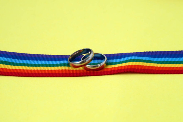 pair of white golden wedding rings on LGBTQ rainbow ribbon on yellow background - Φωτογραφία, εικόνα