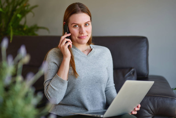 Femme heureuse assise sur un canapé avec ordinateur portable et parlant au téléphone à la maison
. - Photo, image