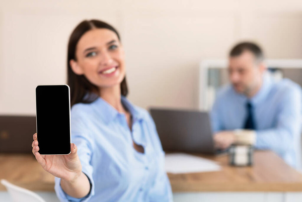 Деловая женщина показывает смартфон с пустым экраном, сидя с коллегой в офисе
 - Фото, изображение