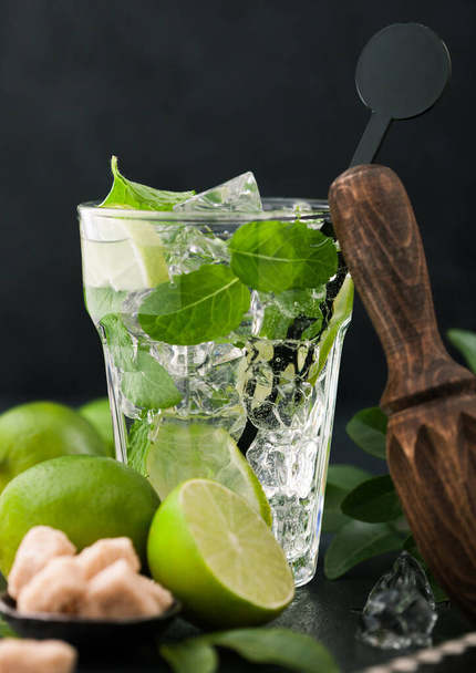 Glass of Mojito cocktail with ice cubes mint and lime on black board with spoon and fresh limes and cane sugar with wooden squeezer and steel shaker with ice.Best summer cocktail - Foto, Bild
