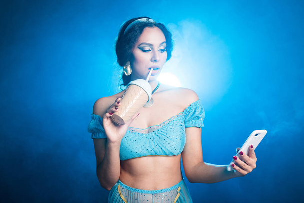 Magic, cosplay and fairy tale concept - Portrait of a young woman in the image of an Eastern fairy Princess holds smartphone and shaker on blue background. - Foto, Imagem