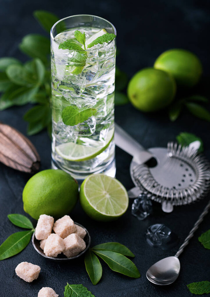 Highball glass of Mojito cocktail with ice cubes,mint and lime on black board with spoon and strainer and fresh limes with cane sugar and wooden squeezer and black shaker. Best party drink. - Foto, afbeelding