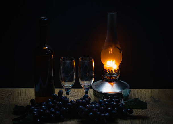 a bottle of red wine and two glasses against a background of black grapes in the dim light from a kerosene lamp - Foto, Imagem