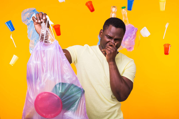 Environmental pollution, plastic recycling problem and waste disposal concept - angry african american man holding garbage bag on yellow background. He is feel smell of trash. - Photo, Image