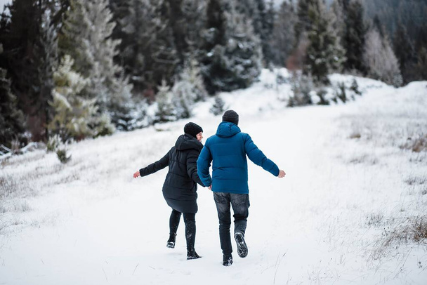 Beautiful young couple running in the mountains holding hands between Christmas trees in winter. Snow. Love of young couple. Free space for text is available. - Fotoğraf, Görsel