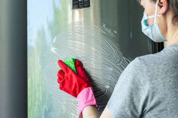 Koruyucu maskeli beyaz bir kadın, temiz süngerli kırmızı lastik eldivenler paslanmaz çelik buzdolabının kapısına sabun köpüğü sürüyor. Konsept: Temizlik, temizlik servisi. Seçici odak. - Fotoğraf, Görsel