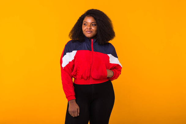 Amusante fille afro-américaine frisée vêtue de la veste de sport rouge dans le studio sur fond jaune avec copyspace. Mode et concept de personnes
. - Photo, image
