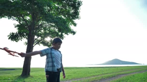 幸せなアジアの子供の男の子は、夕方に太陽の光がある自然の中で、木々や草原を屋外でシミュレートする飛行機を投げている。幸福の休日スローモーション - 映像、動画
