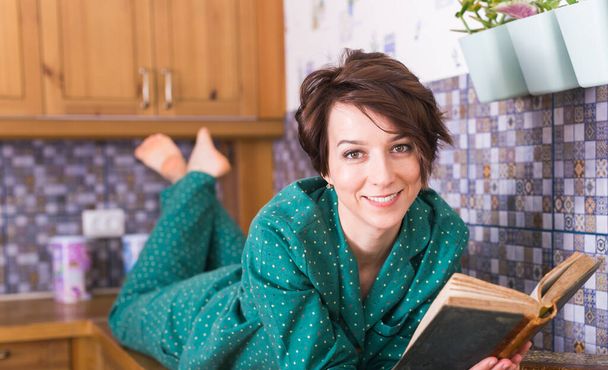Mujer feliz lleva pijama y relajarse en casa y leer un libro. Quédate en casa. Concepto de coronavirus pandémico de cuarentena
. - Foto, Imagen