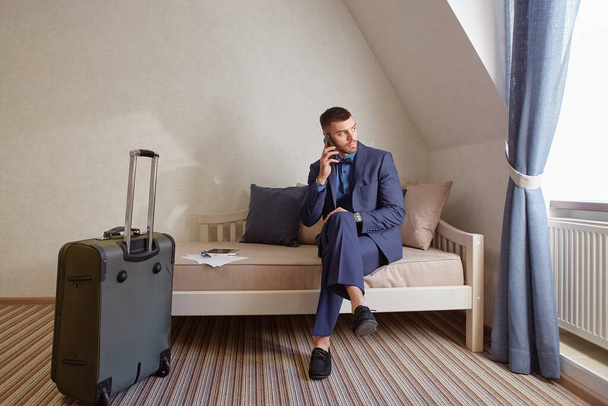 Молодий бізнесмен, використовуючи телефон, сидячи в затишному готельному номері з валізою
. - Фото, зображення