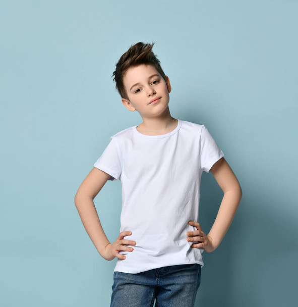Teenager with stylish haircut, dressed in white t-shirt. Smiling, folded his hands and looking up, posing against blue background - Foto, afbeelding
