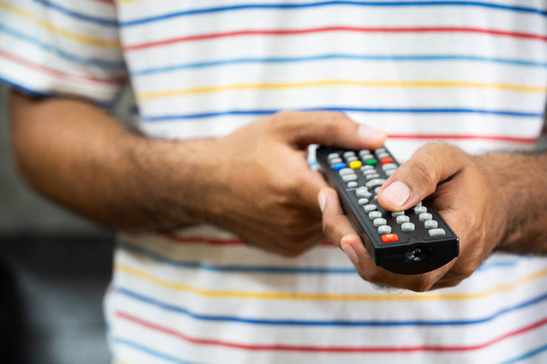 Young man holding television remote control. Hands pointing to tv set and turning it on or off select channel watching tv on his sofa at home in the living room relax. - Photo, Image