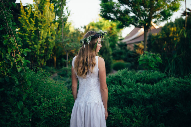 girl in a white dress stands with a wreath on her head against a background of trees and sky - Фото, изображение