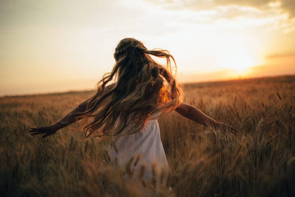 dziewczyna w białej sukience z rozkładanymi ramionami stoi w polu pszenicy na tle zachodu słońca - Zdjęcie, obraz