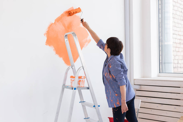 Kaunis keski-ikäinen nainen maalaamassa seinää. Remontointi, remontointi ja korjaus käsite. - Valokuva, kuva