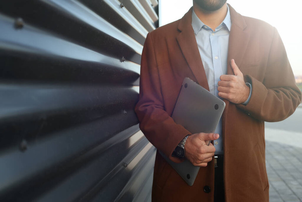 A young guy in a coat and with a laptop posing against a metal wall - Фото, изображение