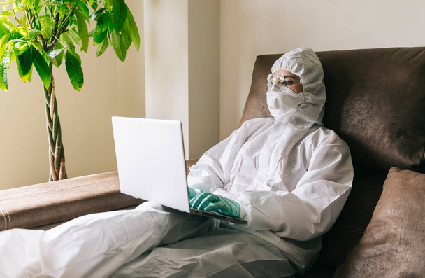 A woman works from home with the laptop, digital tablet and mobile phone in the living room, equipped with security measures against a virus prevention with a safety suit, glasses, mask and latex gloves. - Foto, Imagem
