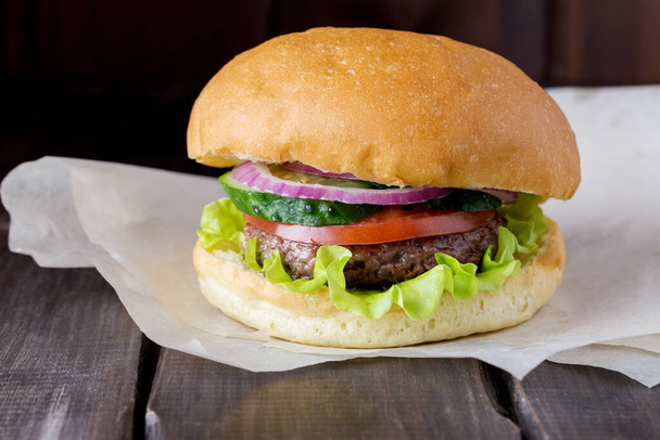 Big tasty burger at craft paper and dark wood background. Fresh bun with red onion, cucumber, tomatoes, beef and green salad. - Φωτογραφία, εικόνα