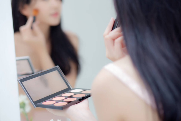 Beleza jovem mulher asiática feliz e sorriso com rosto olhando espelho aplicando maquiagem com escova de bochecha no quarto, bonito de menina segurando escova make-up no facial, cuidados com a pele e conceito cosmético. - Foto, Imagem
