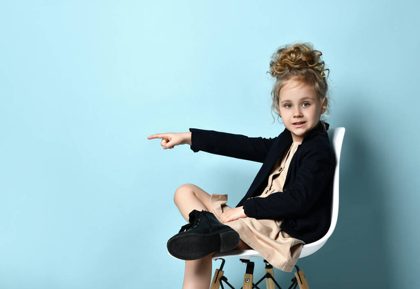 Little model in beige dress, black jacket and boots. She pointing at something, sitting cross-legged against blue background - Photo, image