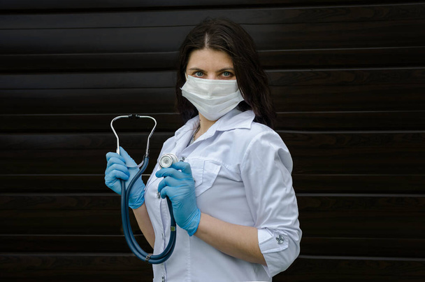 Una joven doctora con una máscara y guantes azules con un estetoscopio en las manos mira a la cámara
. - Foto, imagen