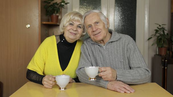 Ευτυχισμένο ζευγάρι παππούδων κάθονται στο τραπέζι στο σπίτι και απολαμβάνουν το τσάι μαζί. - Φωτογραφία, εικόνα