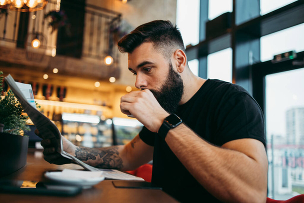 Young adult hipster man with a long beard sitting in modern cafe bar, drinking coffee and reading newspapers. - Fotografie, Obrázek