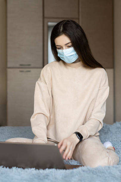 Yatağın üzerinde koruyucu mavi tıp maskeli genç bir kız öğrenci dizüstü bilgisayarla çalışıyor, online eğitim alışverişi öğreniyor, karantina sırasında evde çalışıyor. - Fotoğraf, Görsel